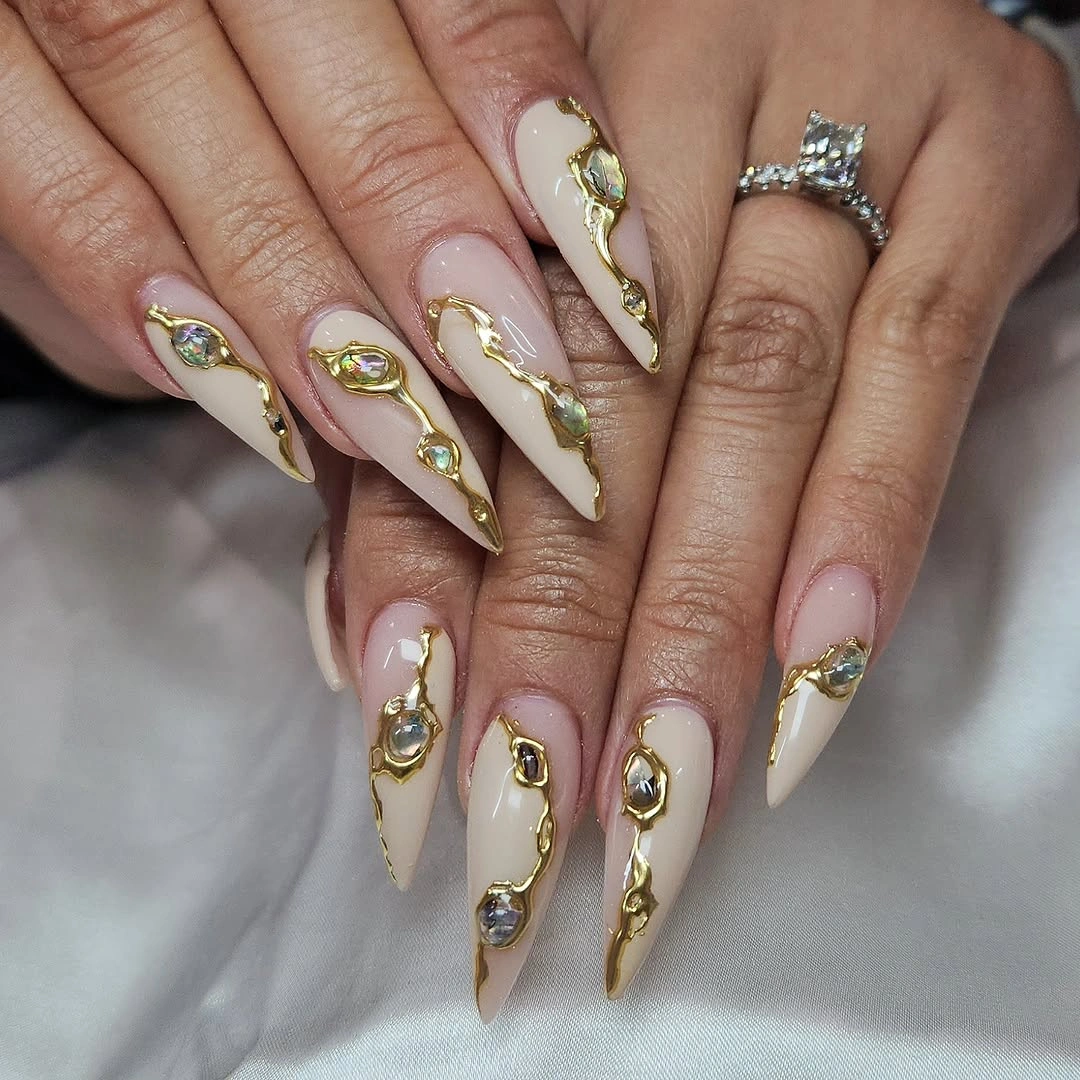 Long ivory and nude pointed nails with raised-looking gold curves and iridescent accents
